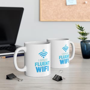 May include: Two white ceramic mugs with the text "I Speak Fluent Wifi" in blue. Each mug features a blue wifi symbol with steam. The mugs are on a light-coloured surface with a laptop and office supplies in the background.