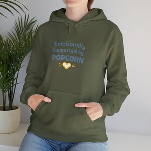 Puede incluir: Sudadera con capucha verde oliva con bolsillo delantero y capucha con cordón. La sudadera tiene el texto "Emotionally Supported by POPCORN" en azul, con un gráfico de corazón de palomitas de maíz. El modelo lleva vaqueros azules.