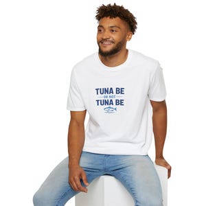 Peut inclure: T-shirt blanc avec l'inscription bleue marine "TUNA BE or not TUNA BE" et un dessin de poisson. La personne porte un jean bleu.