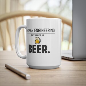 Puede incluir: Taza de cerámica blanca con el texto "GERMAN ENGINEERING. BUT MAKE IT BEER." en negro, con un pequeño gráfico de jarra de cerveza. La taza está sobre una superficie de madera con un bolígrafo y un portátil en el fondo.