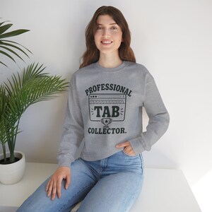 Könnte beinhalten: Ein graumeliertes Langarm-Sweatshirt mit Rundhalsausschnitt und einem schwarzen Grafikdruck eines Browserfensters, dem Text "PROFESSIONAL TAB COLLECTOR." sowie einem kleinen Trophäensymbol auf der Brust, kombiniert mit einer hellblauen Jeans.