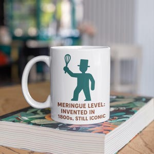 Könnte beinhalten: Weiße Keramik-Tasse mit einer türkisfarbenen Silhouette einer Person mit Schneebesen und Zylinder. Text: MERINGUE LEVEL: INVENTED IN 1800s, STILL ICONIC. Die Tasse steht auf einem Buch mit farbenfrohem Einband.