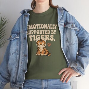 May include: An olive green t-shirt featuring the text "Emotionally Supported by Tigers" and a cartoon tiger. The tiger is orange and black with a smiling face and a small red heart. The shirt is made of a soft material.