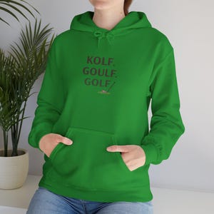 Può includere: Una felpa con cappuccio verde con tasca frontale e coulisse. La felpa presenta le parole "KOLF. GOULF. GOLF/" in un font rosso, insieme a una piccola grafica di mazza e pallina da golf.