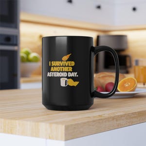 May include: Black ceramic coffee mug with a curved handle. The mug features the text "I survived another asteroid day" in yellow and white, along with a graphic of a comet and a coffee cup.