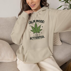 May include: A beige hooded sweatshirt featuring a graphic print of a smiling green cannabis leaf above a power button icon. The text above the leaf reads CALM MODE: and the text below the icon reads ACTIVATED. The hoodie has long sleeves and a front pocket.