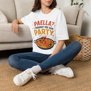 May include: A white short-sleeved t-shirt featuring a graphic of a cartoon shrimp wearing sunglasses inside a paella pan. The text reads PAELLA? I THOUGHT YOU SAID PARTY in red, black, and yellow lettering, accompanied by small confetti-like shapes.