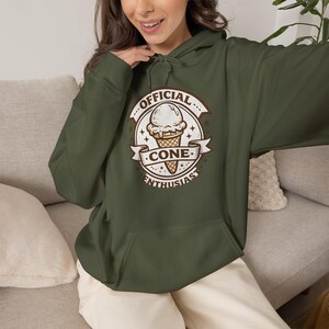 Op de afbeelding: Een olijfgroene hoodie met een grafische print van een ijshoorntje in een cirkelvormig embleem. De tekst luidt OFFICIAL CONE ENTHUSIAST in bruine letters. De hoodie is voorzien van een kangoeroezak aan de voorzijde en bijpassende trekkoorden.