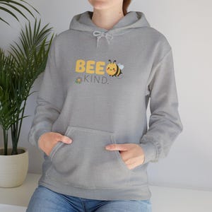 Peut inclure: Un sweat à capuche gris avec une poche avant et un cordon de serrage. Le sweat-shirt affiche le texte "BEE KIND" en jaune, avec un dessin d'abeille et de fleur. Il a des manches longues et une coupe décontractée.