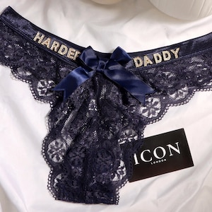 May include: Dark navy blue lace underwear featuring a central satin bow and rhinestone lettering that reads HARDER DADDY across the waistband. A small black rectangular tag with the white text ICON LONDON is tucked into the lace fabric.