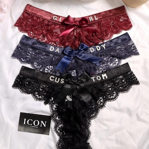 May include: Three pairs of lace underwear in burgundy, navy blue, and black. Each pair features a satin bow at the center and rhinestone lettering on the waistband reading GIRL, DADDY, and CUSTOM. A black tag with the white text ICON LONDON is visible.