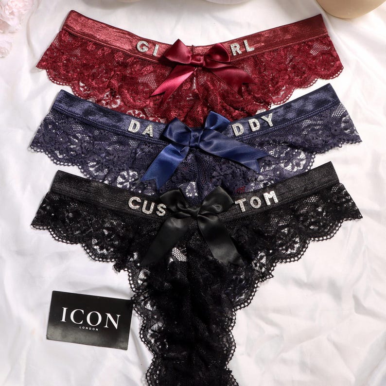 May include: Three pairs of lace underwear in burgundy, navy blue, and black. Each pair features a satin bow at the center and rhinestone lettering on the waistband reading GIRL, DADDY, and CUSTOM. A black tag with the white text ICON LONDON is visible.