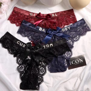 May include: Three pairs of lace underwear in burgundy, navy blue, and black. Each pair features a satin bow at the center front and rhinestone lettering on the waistband reading GIRL, DADDY, and CUSTOM. A small black card with ICON LONDON text is included.