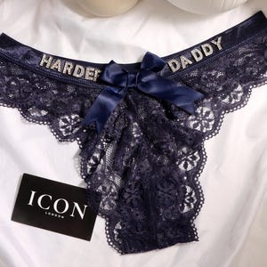 May include: Navy blue lace thong with the words "HARDER DADDY" in silver rhinestones. A navy satin bow is centered on the front. The thong has a scalloped lace trim. A black card with "ICON LONDON" is in the lower left.