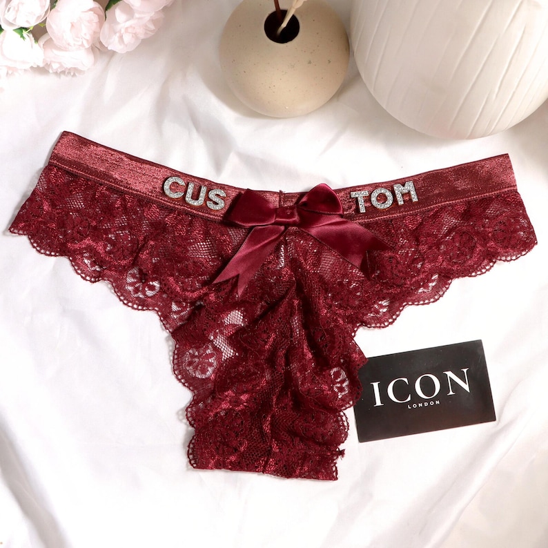 May include: Deep burgundy lace underwear featuring a satin waistband with the word CUSTOM spelled out in silver rhinestone letters. A matching burgundy satin bow is centered on the front. A black rectangular card with the white text ICON LONDON is placed next to the garment.