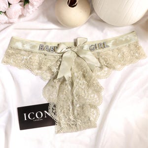 May include: A pair of light beige lace underwear featuring a shimmering elastic waistband with the text BABY GIRL in silver rhinestones. A matching satin ribbon bow is centered at the front. A black rectangular tag displays the text ICON LONDON.