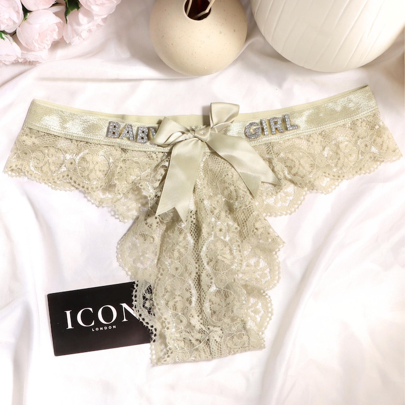 May include: A pair of light beige lace underwear featuring a shimmering elastic waistband with the text BABY GIRL in silver rhinestones. A matching satin ribbon bow is centered at the front. A black rectangular tag displays the text ICON LONDON.
