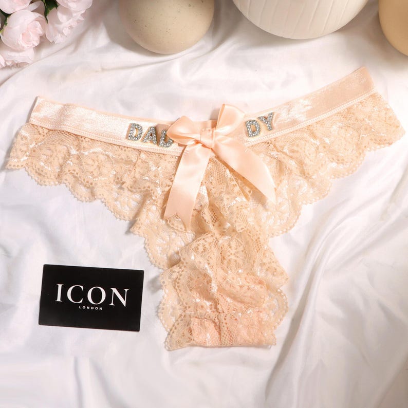 May include: A peach-colored lace panty featuring a satin waistband with the word DADDY spelled out in silver rhinestones. A matching satin bow is centered on the front. A black rectangular card with the white text ICON LONDON is included.