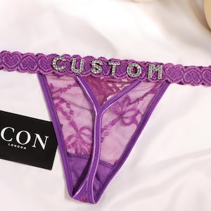 Peut inclure: Un string en dentelle violette doté d'une ceinture à motif cœurs et du mot CUSTOM écrit en lettres strass argentées. Une petite carte rectangulaire noire portant le texte blanc ICON LONDON est placée à côté du vêtement.