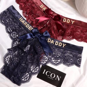 Pode incluir: Três pares de roupa interior de renda em azul marinho e bordô, com as palavras "HARDER" e "DADDY" em letras douradas. Cada par tem um laço de cetim. Também está presente um cartão preto e branco com o texto "ICON LONDON".