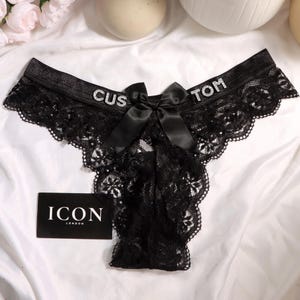May include: Black lace underwear featuring a satin bow at the center front and rhinestone lettering on the waistband that reads CUSTOM. A small black rectangular tag with the white text ICON LONDON is positioned near the bottom left corner.
