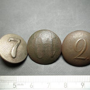 Könnte beinhalten: Drei antike Metallknöpfe mit den geprägten Zahlen 7, 11 und 2. Die Knöpfe sind kreisförmig und leicht gewölbt. Ein Lineal befindet sich am unteren Rand, mit Maßen in Zentimetern.