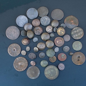 May include: A collection of assorted vintage metal buttons in various sizes and finishes, including brass, copper, and silver tones. The buttons feature diverse textures, engraved patterns, and some display embossed numbers or military-style insignias.