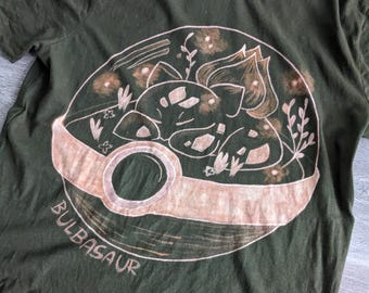 May include: A green t-shirt with a bleach-dyed design of a Pokemon character, Bulbasaur, inside a Pokeball. The design is in shades of brown and white.