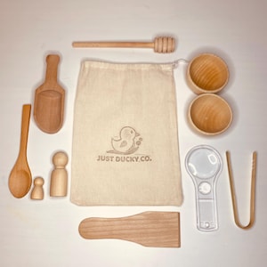 May include: A collection of wooden toys and tools, including a scoop, spoon, bowls, tongs, a magnifying glass, and a honey dipper. A canvas bag with the text "JUST DUCKY CO." is also included. The items are arranged on a white surface.