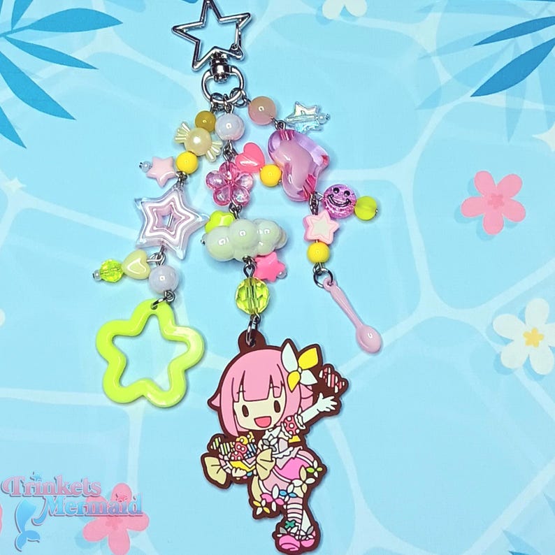 May include: A whimsical keychain featuring a pink anime-style character charm, surrounded by star, flower, and heart-shaped beads in pastel colors. The keychain includes a star-shaped clasp and a variety of decorative elements.