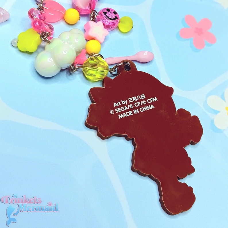 May include: A brown charm with a character design, attached to a beaded keychain with pink, yellow, and white beads. The charm has text that reads "Art by 三月八日 &copy; SEGA/CP/&copy; CFM MADE IN CHINA". The background is light blue with flower designs.
