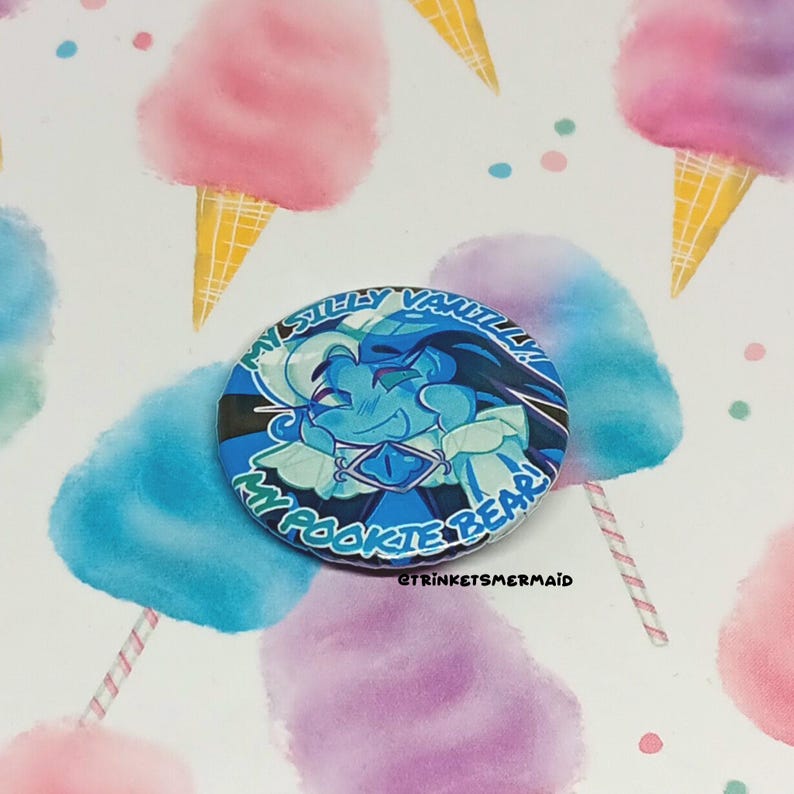 May include: A blue and white button with a cartoon character with blue skin and a white collar. The text on the button reads "My Silly Vanilla Rookie Bear!"