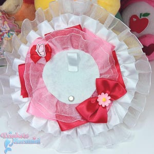 May include: A decorative pin in white, pink, and red. It has a white felt backing, a red bow with a pink flower, and a small red bow with a butterfly. The pin has a layered, ruffled edge. The pin is a decorative accessory.