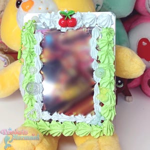 May include: A rectangular mirror with a decorative frame. The frame is adorned with white and light green frosting-like embellishments, small flowers, and a pair of red cherries. The mirror is surrounded by plush toys.