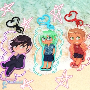 Puede incluir: Tres llaveros de personajes de estilo anime con clips de diferentes colores. Un personaje viste un traje, otro una camisa hawaiana y el tercero una sudadera con capucha roja. El fondo tiene formas de estrellas y corazones.