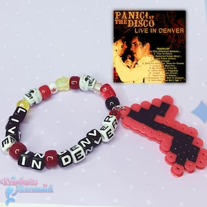 May include: A beaded bracelet spelling "LIVE IN DENVER" with black and white beads, interspersed with red and skull beads. A red and black cross charm is attached. A CD case is visible in the background.
