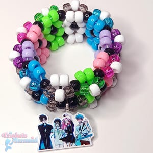 May include: A multi-layered cuff bracelet made of colorful plastic pony beads in shades of white, black, neon green, bright blue, lavender, and hot pink. A flat, rectangular charm featuring an anime-style illustration of four characters is attached.