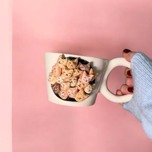 May include: A white ceramic mug with a large, round handle. The mug features a 3D relief of numerous cat faces in shades of tan, brown, and cream, clustered together on a black background. The mug is held against a pink backdrop.