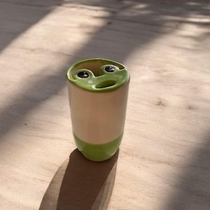 May include: A ceramic tumbler with a frog face design. The top rim and base are green, with a beige body. The frog's eyes are black, and the mouth is open. The tumbler is cylindrical and sits on a wooden surface.