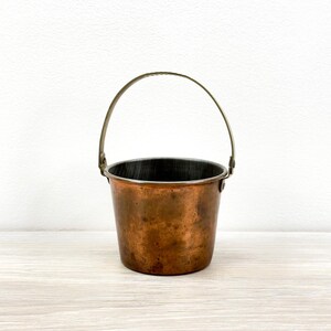 May include: A small, round copper bucket with a weathered, dark metallic patina and a smooth, dark interior. The bucket features a thin, arched metal handle attached to the sides with circular rivets, creating a simple, functional design.