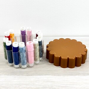 May include: A collection of sewing thread spools in various colors including blue, pink, green, and yellow, housed in a clear plastic circular organizer, positioned next to a brown, scalloped-edge plastic lid for the thread storage container.
