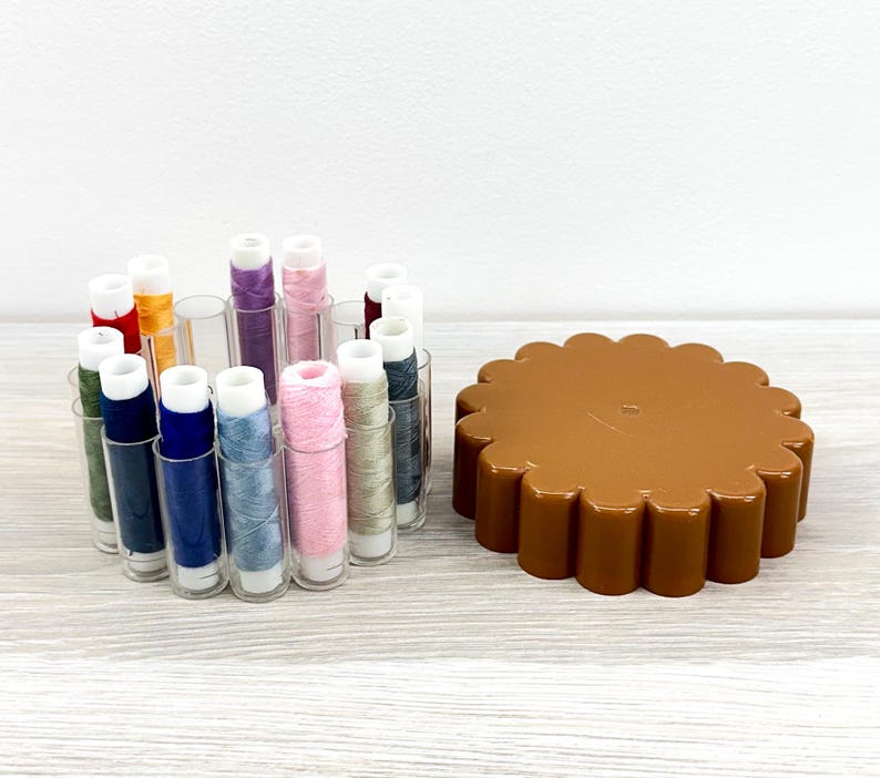May include: A collection of sewing thread spools in various colors including blue, pink, green, and yellow, housed in a clear plastic circular organizer, positioned next to a brown, scalloped-edge plastic lid for the thread storage container.