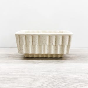 May include: A square ceramic bowl with a cream-colored glaze and dark speckles. The exterior features a repeating vertical ribbed pattern around the sides, and the base is slightly recessed, creating a tiered effect for this small kitchen vessel.