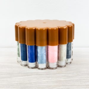 May include: A circular brown plastic storage container featuring multiple vertical compartments, each holding a spool of sewing thread in various colors including blue, pink, grey, and green, arranged in a radial pattern with a matching lid.