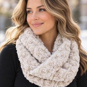 May include: A cream-colored infinity scarf featuring a textured, swirling rose-like pattern throughout the fabric. The scarf is worn around the neck over a solid black long-sleeved shirt, providing a soft, plush appearance with a consistent floral-inspired design.