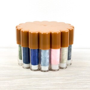 May include: A circular plastic storage container with a scalloped edge lid in a tan color, holding multiple vertical clear tubes filled with various colors of sewing thread including blue, pink, green, and grey, arranged in a compact cluster.