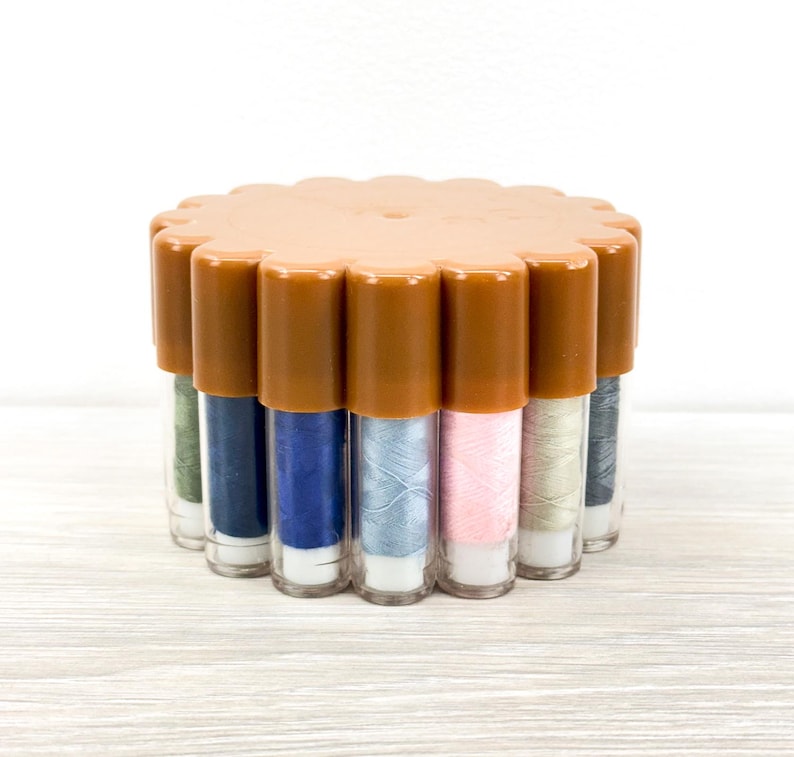 May include: A circular plastic storage container with a scalloped edge lid in a tan color, holding multiple vertical clear tubes filled with various colors of sewing thread including blue, pink, green, and grey, arranged in a compact cluster.