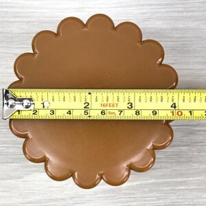 May include: A brown, circular object with a scalloped edge, measuring approximately 4.5 inches in diameter as indicated by the yellow measuring tape placed horizontally across the center of the item.