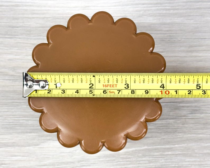 May include: A brown, circular object with a scalloped edge, measuring approximately 4.5 inches in diameter as indicated by the yellow measuring tape placed horizontally across the center of the item.