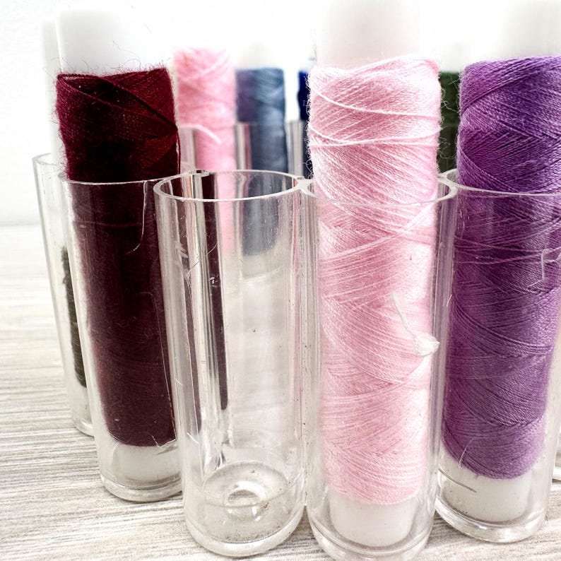 May include: A collection of cylindrical spools of sewing thread in various colors including deep burgundy, light pink, dark blue, and purple, each held upright in individual clear plastic tubes arranged in a cluster for organized storage.